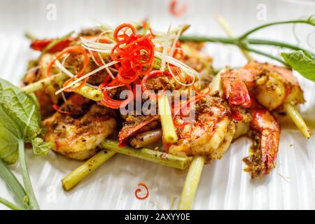 Grilled king prawns in spicy sauce on lemongrass skewers. Decorated with red chili. On a white ceramic plate. Stock Photo