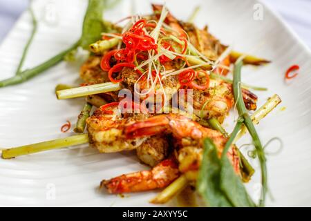 Grilled king prawns in spicy sauce on lemongrass skewers. Decorated with red chili. On a white ceramic plate. Stock Photo