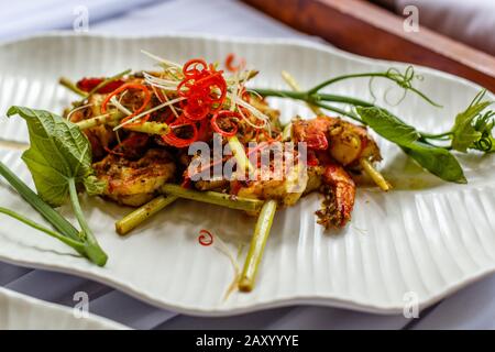 Grilled king prawns in spicy sauce on lemongrass skewers. Decorated with red chili. On a white ceramic plate. Stock Photo