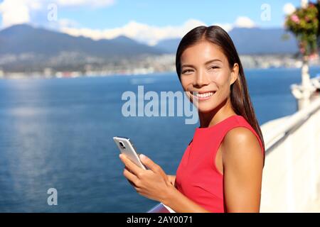 Happy asian woman using phone app in vancouver city harbour on summer day. Beautiful landscape of canadian nature river and mountains. Businesswoman on smartphone lifestyle. Stock Photo