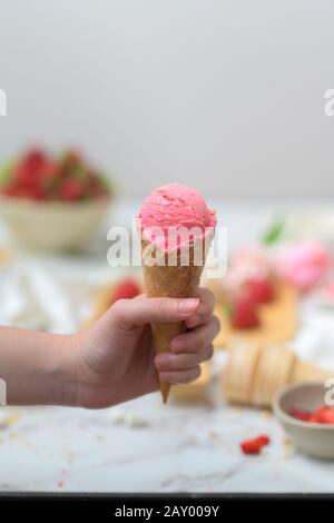 Cropped shot of a girl holding strawberry ice-cream cone with blurred strawberry and topping on marble table background Stock Photo