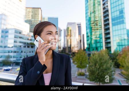 Smart phone woman calling on mobile phone in city business district. Confident young business woman talking on smartphone smiling happy wearing suit jacket outdoors. Urban female professional in 20s. Stock Photo