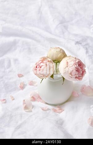 Crumpled white sheets of bed with small ceramic vase with pink rose flowers and tender petals on it, copy space. Valentine's day greeting card. Stock Photo