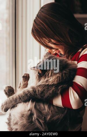 Smiling girl standing by a window cuddling her cat Stock Photo