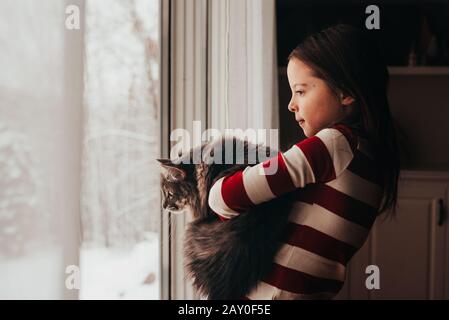 Girl standing by a window cuddling her cat Stock Photo