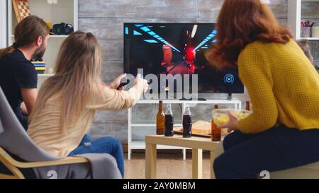 Back view of blonde woman playing shooter games while her friends are eating pizza. Playing games with wireless controller. Stock Photo