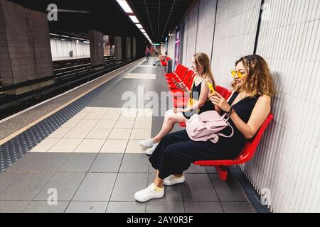 Two girl friends reading smartphon in metro while waiting for the train. Social media and internet addiction concept Stock Photo