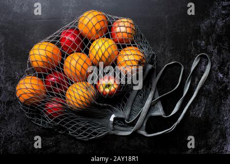 High angle close up of red apples and oranges in grey net bag on black background. Stock Photo