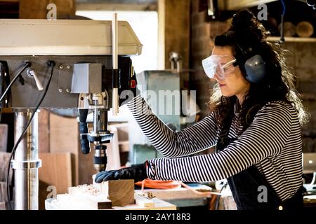 Woman with long brown hair wearing dungarees, safety glasses and ear protectors standing in wood workshop, using electric drill. Stock Photo