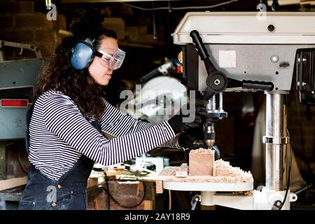 Woman with long brown hair wearing dungarees, safety glasses and ear protectors standing in wood workshop, using electric drill. Stock Photo