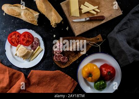 High angle close up of a selection of cheeses, tomatoes, salami and French baguette on white enamel plates on black background. Stock Photo