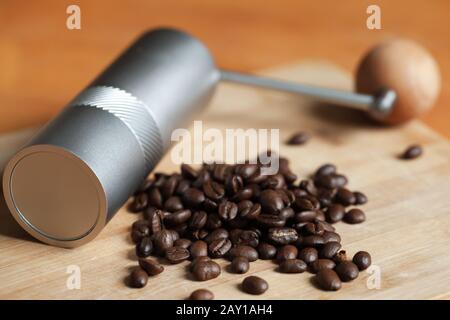 Shiny metallic manual coffee grinder and dark roasted coffee beans are on wooden desk, close-up photo with soft selective focus Stock Photo