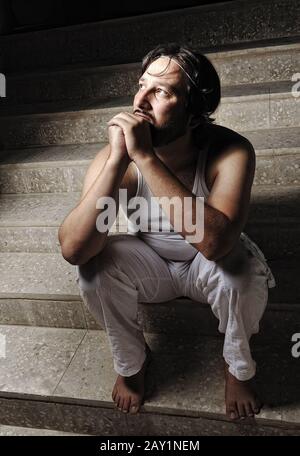 Thoughtful man on black background in low key, sitting on stairs Stock Photo