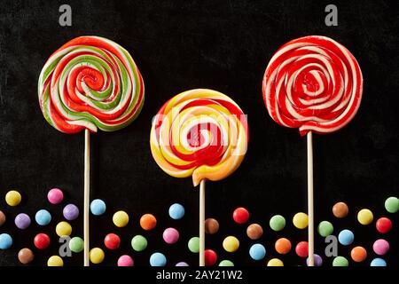 Three brightly colored red, green and yellow lollipops with a border of sugar-coated chocolate candy on a black background Stock Photo