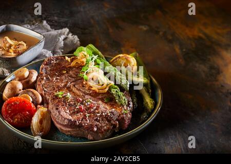 Seasoned thick grilled beef steak with fresh green spring asparagus, roasted onion, tomato and mushrooms on a dark wooden counter Stock Photo