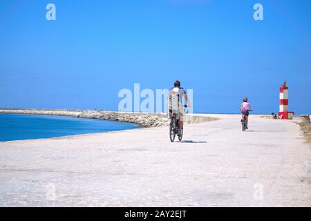 Couple and their child riding bicycles towards a small light house on a warm summers day next to a beautiful blue beach.