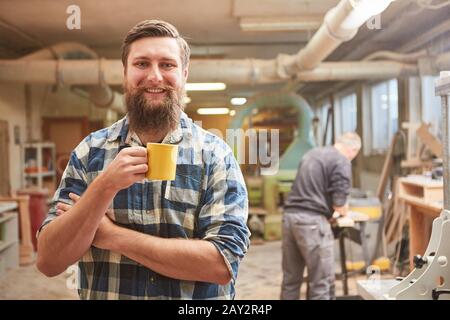 Smiling hipster carpenter with beard takes coffee break in his joinery Stock Photo