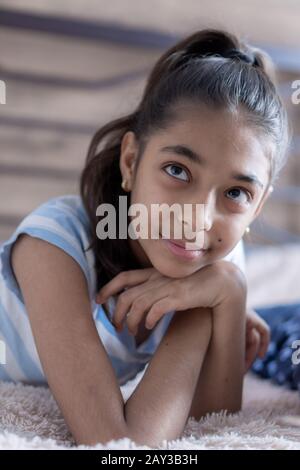 Closeup portrait of a young swarthy girl on a bed in the sun. Persian black-haired girl cute looks lying on the bed holding a smartphone. Stock Photo