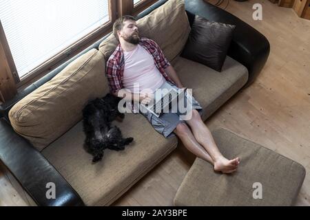 Bearded man with a laptop fell asleep on a soft sofa, next to him funny, belly up, lies his dog Schnauzer, in natural light. Stock Photo