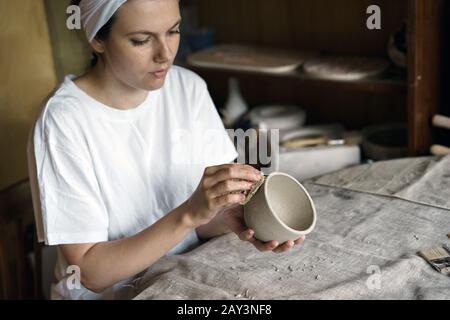 Beautiful woman in a creative workshop sits at a table and wipes a clay bowl Stock Photo