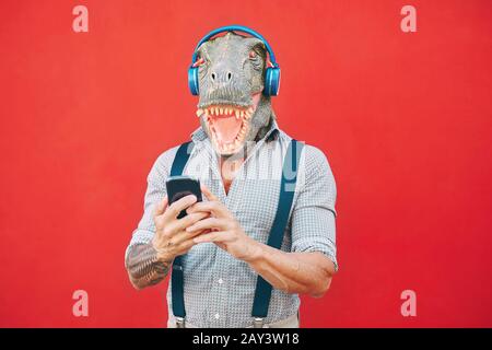 Senior fashion man wearing t-rex mask using mobile smartphone listening music with headphones - Crazy bizarre hipster guy having fun Stock Photo