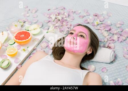 Beautiful young woman is getting facial clay mask at spa, lying with cucumbers on eyes Stock Photo