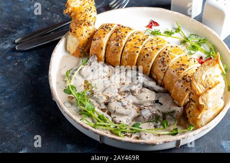 Puff pastry roll stuffed with mushrooms. Nearby are champignons in sour cream, sliced. Served with microgreen and sun-dried tomatoes. On a dark backgr Stock Photo