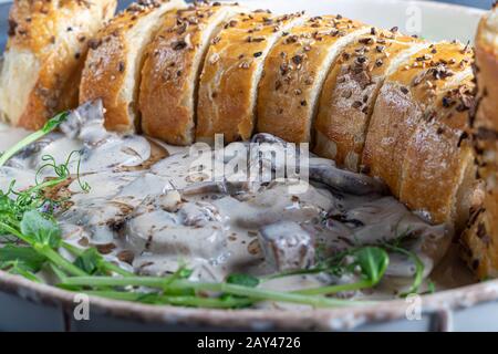 Puff pastry roll stuffed with mushrooms. Nearby are champignons in sour cream, sliced. Served with microgreen and sun-dried tomatoes. On a dark backgr Stock Photo