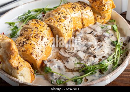 Puff pastry roll stuffed with mushrooms. Nearby are champignons in sour cream, sliced. Served with microgreen and sun-dried tomatoes. On a dark backgr Stock Photo