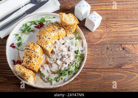 Puff pastry roll stuffed with mushrooms. Nearby are champignons in sour cream, sliced. Served with microgreen and sun-dried tomatoes. On a dark backgr Stock Photo
