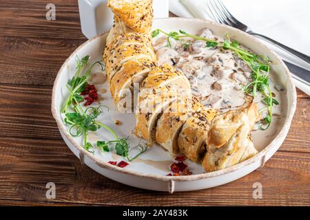 Puff pastry roll stuffed with mushrooms. Nearby are champignons in sour cream, sliced. Served with microgreen and sun-dried tomatoes. On a dark backgr Stock Photo