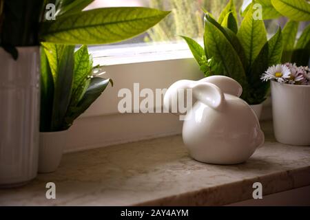 White porcelain easter bunny on windowsill with green leaves deco and blooming daisies in flowerpot. Waiting for spring, background with copy space, Stock Photo