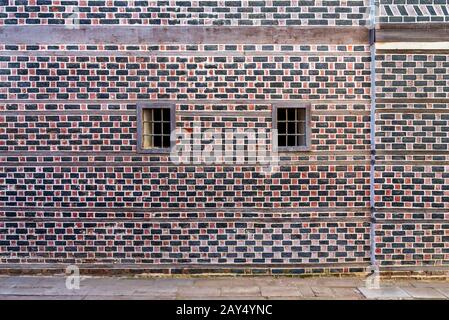 Wall with black and red bricks with white seam and two small adjacent windows with metal bars on cobblestone road Stock Photo