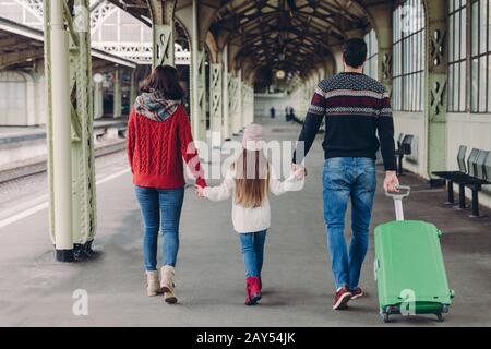 Back view of friendly family hold hands, carry suitcase, going to have voyage trip, pose on railway station platform. Young mother and father, their s Stock Photo