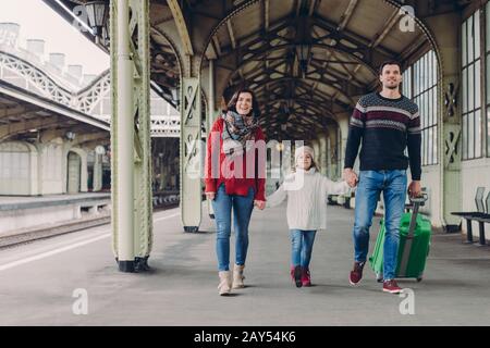Shot of happy family going to have nice trip during holidays, carry bag, walk on railways station platform, being in good mood. Father, mother and chi Stock Photo