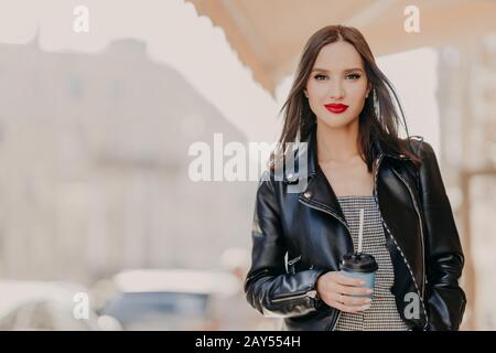 Outdoor shot of attractive millennial female model with makeup, dressed in stylish leather jacket, drinks coffee from disposable cup, wanders around c Stock Photo