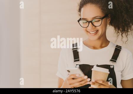 Adorable teeange girl with curly hair, checks news feed, messages in social networks, uses free internet connection, wears optical glasses, casual whi Stock Photo