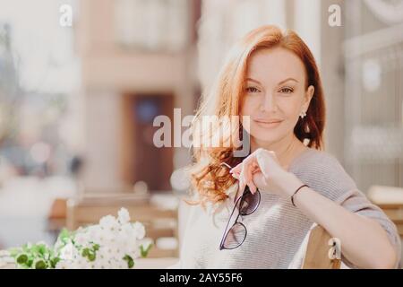 Horizontal shot of attractive pleased red haired woman sits on bench, enjoys sunny day, holds sunglasses, breathes fresh air, has make up, poses over Stock Photo