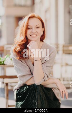 Cheerful attractive red haired young woman has positive expression, keeps hand under chin, wears fashionable attire, looks direclty at camera, poses o Stock Photo