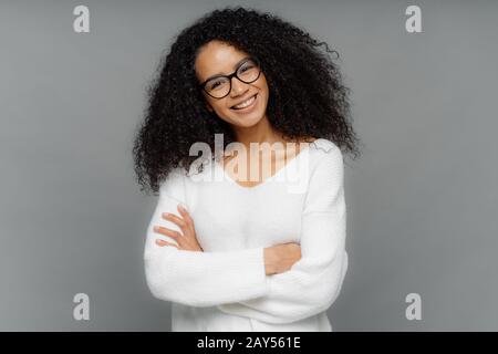 Half length shot of friendly looking African American woman tilts head, has dark crisp bushy hair, keeps arms folded over chest, wears casual white sw Stock Photo
