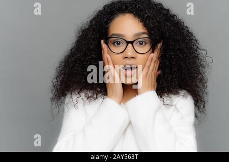 Close up portrait of shocked concerned young woman with dark skin and curly hair, hears terrifying news, keeps hands on cheeks, wears spectacles and w Stock Photo