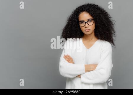 Self confident dark skinned ethnic female keeps hands crossed over chest, looks seriously at camera, wears optical spectacles and white sweater, stand Stock Photo