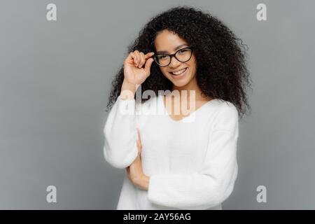 Portrait of happy woman with curly hair, kepes hands on frame of glasses, has gentle smile, wears white jumper, isolated over grey background. Copy sp Stock Photo