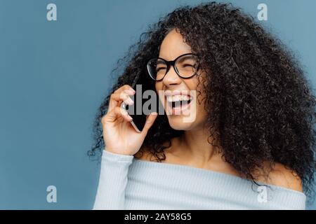 Joyful female has Afro hair, laughs happily, talks via cellular, discusses something funny with friend, smiles broadly, shows white teeth, isolated ov Stock Photo