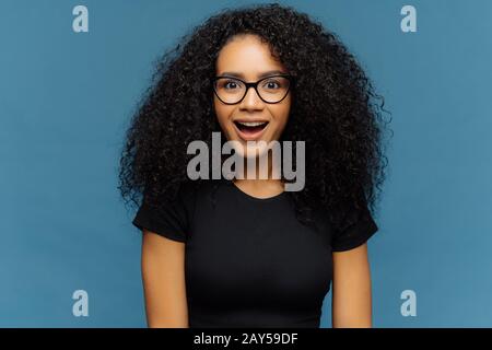 Pleasantly surprised dark skinned woman keeps jaw dropped, gazes with interest, has curly hair, wears black t shirt, models against blue wall. Afro Am