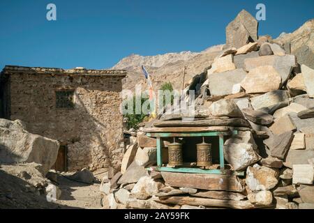 Street flanked by religious symbols, houses, prayer flags, and stone boulder, with Himalayas as backdrop on sunny day in Nako, Himachal Pradesh, India. Stock Photo