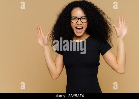 Half length shot of pleased overjoyed woman dances and moves positively, raises palms, exclaims with happiness, wears black t shirt, spectacles, isola Stock Photo