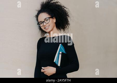 Positive curly African American student carries notepad and diary, wears black clothes, transparent glasses, enjoys studying in university, isolated o Stock Photo