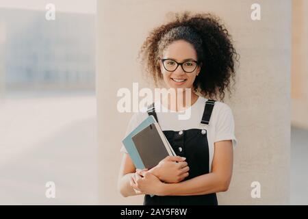 Adorable curly haired female student wears white casual t shirt and overalls, holds notepad or textbook, looks through transparent spectacles, stands Stock Photo