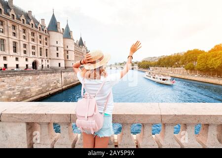 A girl tourist in a hat standing on a bridge and waving to a river cruise sailing by Seine river in Paris Stock Photo